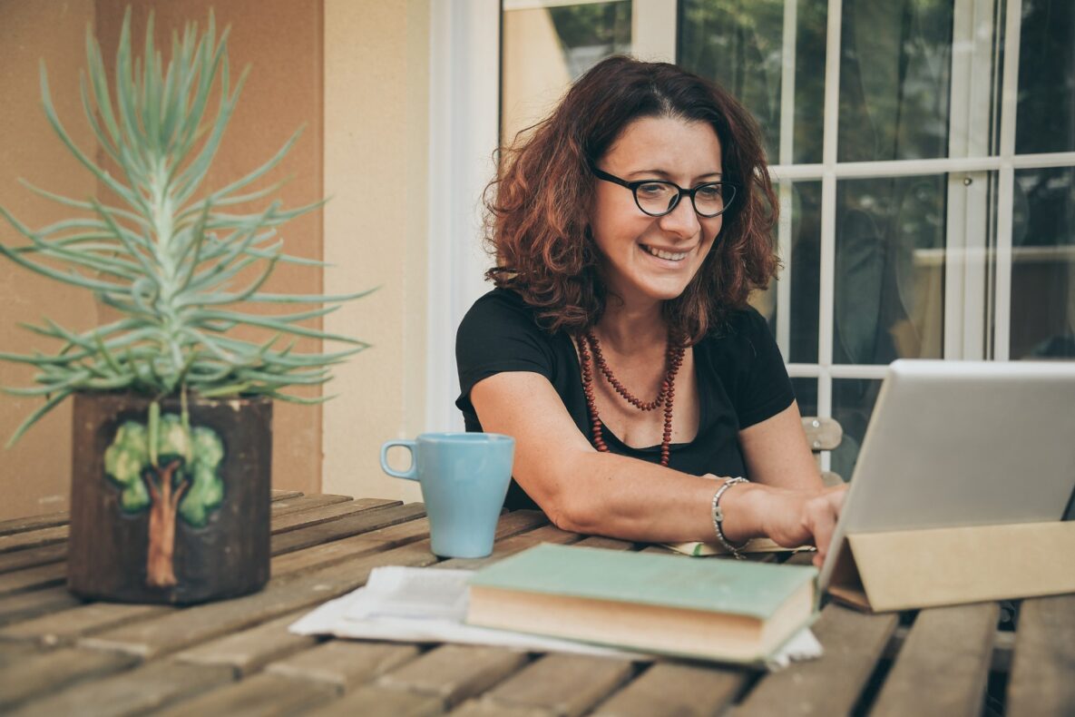 Middle aged female studying at home with books, newspaper and digital tablet pad. Woman reading a book and watching video online on new tech device. Education, modern lifestyle and leisure concept.