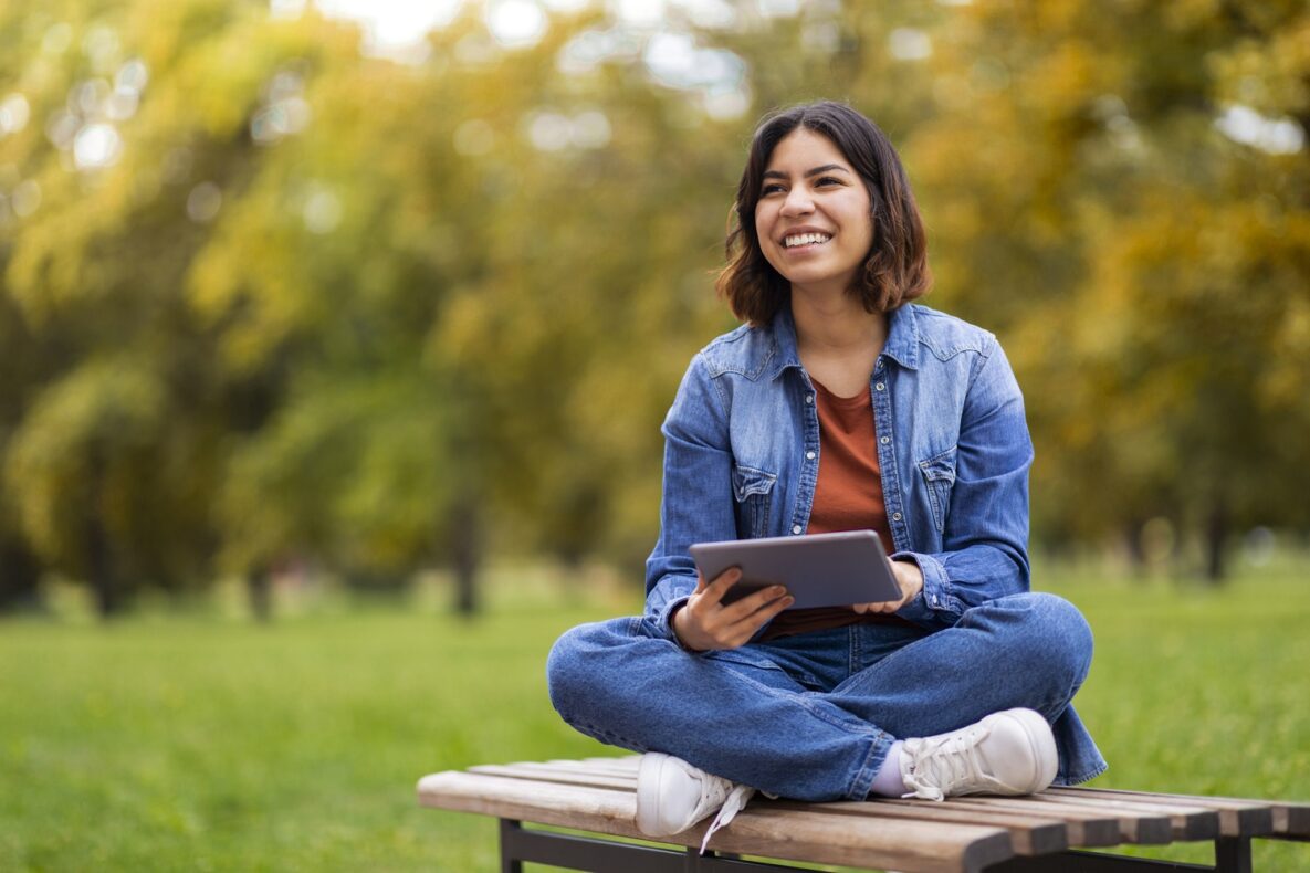 Happy Beautiful Young Arab Woman Relaxing With Digital Tablet Outdoors, Cheerful Middle Eastern Female Sitting on Bench In Park And Using Modern Gadget, Browsing Internet Or Checking Social Media
